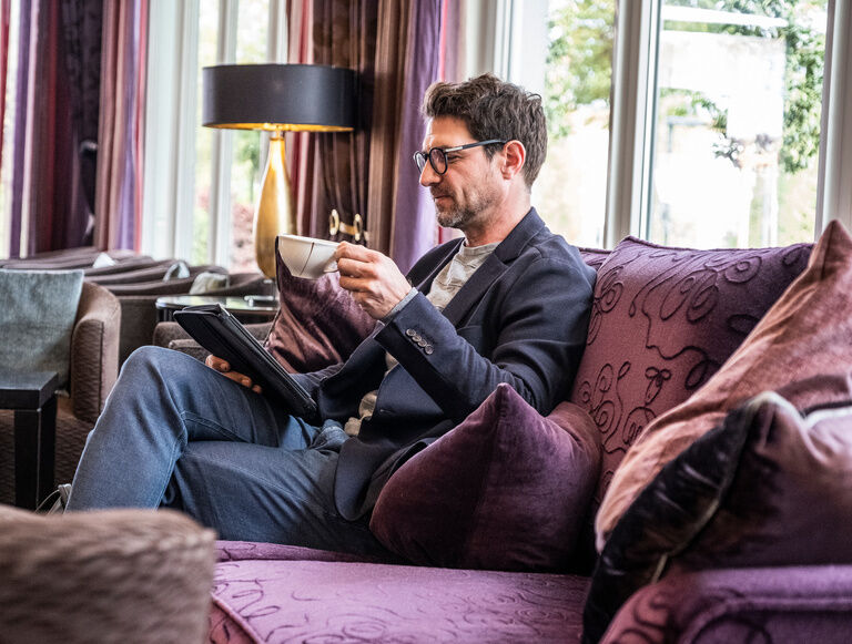 Ein Mann sitzt auf einem gemütlichen lila Sofa in der Lounge des Hotel Holzapfel und liest ein Buch.