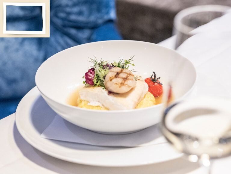 Ein sorgfältig arrangiertes Gericht in einer weißen Schüssel mit einem Glas Wein auf einem Tisch im Wellnesshotel Holzapfel.