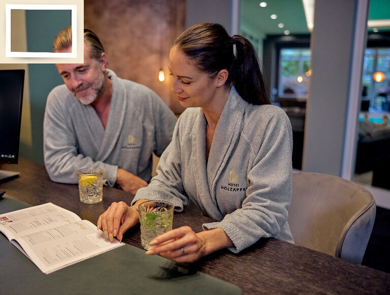 Ein Paar am Tisch der Rezeption des Gesundheitshotels Holzapfel in Bayern wirkt entspannt und lächelt während des Genusses eines Getränks.